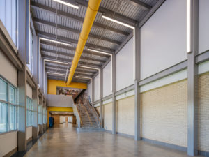 A bright, modern school atrium with a high ceiling, exposed structural beams, and a large central staircase. The space features a mix of polished concrete floors, light brick walls, and a pop of color from a prominent yellow duct and accent wall.