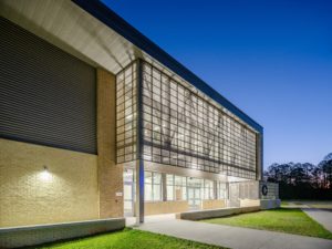 A modern school building entrance at dusk, featuring a contemporary design with a mix of tan brick, gray metal siding, and a prominent, multi-story glass facade that is brightly lit from within.