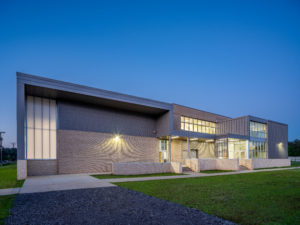 A modern school building exterior at dusk, featuring a contemporary design with a mix of gray brick, metal siding, and large windows. The building has a stepped entrance and is surrounded by a well-maintained lawn under a clear blue sky.