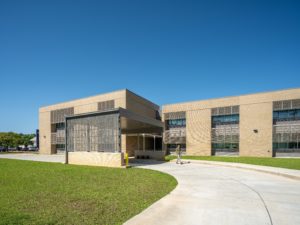 A modern, tan brick school building with a large covered entry area and a prominent, slatted-wood or metal screen facade. The photo is taken on a sunny day with a clear blue sky, showing a curved concrete walkway and a well-maintained grassy lawn.
