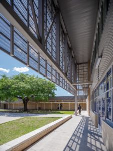 A modern school building's exterior walkway overlooking a central courtyard with a large tree. The walkway features an architectural design with an extended roof, large windows, and a patterned shadow cast on the concrete path.