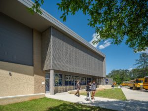 A modern school building exterior on a sunny day, with students walking on a sidewalk. The building features a brick and metal facade with large, screened windows and a sign that reads "Covington." School buses are visible in the background.