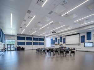 A multi-purpose school room with a polished concrete floor and a large screen for presentations. The modern space features blue acoustic panels on the walls, recessed lighting, a sloped ceiling, and rows of chairs arranged for a meeting or class.