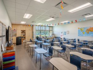 A brightly lit and organized modern classroom with individual desks and chairs, a world map on the wall, and international flag decorations. The room features large windows, built-in cabinetry, and a bulletin board.