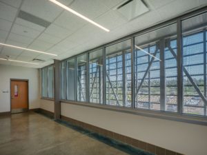 A wide shot of a modern conference room with a long table and chairs, exposed brick walls, and two people seated at the far end. The space features large windows, a colorful purple and black patterned carpet, and a large bench along one wall.