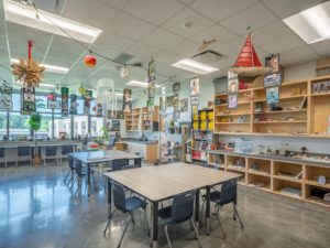 A bright and modern art classroom filled with tables and chairs, a large world map, and international flags hanging from the ceiling. The room is well-lit with natural light and features built-in cabinets and shelving, showcasing a variety of student projects and supplies.