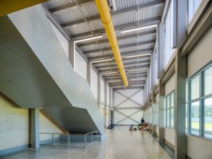 A spacious, modern school hallway with a high ceiling, exposed structural beams, and a prominent yellow ventilation pipe. A large, angular staircase with a perforated metal railing dominates the left, while students are seen sitting on the floor in the distance.