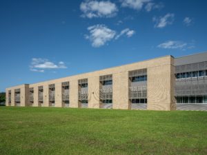 A long, two-story modern school building on a sunny day. The facade features tan brick, large windows with gray metal sunshades, and a clean, contemporary design, surrounded by a green lawn and blue sky.