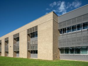A long, modern, two-story school building on a sunny day. The facade is a mix of tan brick and gray metal siding with horizontal louvers over the large windows. The building is surrounded by a lush green lawn under a clear blue sky.