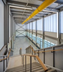 A long shot from the top of an angular staircase looking down a modern school hallway. The space has a high ceiling with exposed structural beams, a bright yellow ventilation pipe running the length of the ceiling, and walls with translucent panels and large windows.