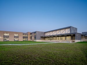 A full exterior shot of a modern school building at dusk, featuring a tan brick facade and a multi-level structure with large, dark-framed windows. The building has a prominent, covered entranceway and is surrounded by a large grassy lawn.