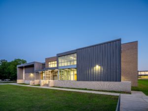 A modern school building exterior at dusk, featuring a contemporary design with a mix of gray brick, vertical metal siding, and large windows. The building has a prominent, multi-level entranceway and is surrounded by a well-maintained lawn under a clear blue sky.