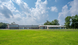 A full exterior view of a modern school building with a distinctive, angular roofline. The low-profile building is set in a large, grassy field with a blue sky and white clouds in the background.