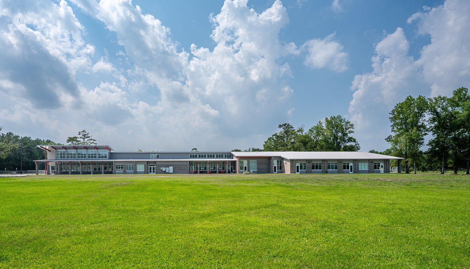 A full exterior view of a modern school building with a distinctive, angular roofline. The low-profile building is set in a large, grassy field with a blue sky and white clouds in the background.
