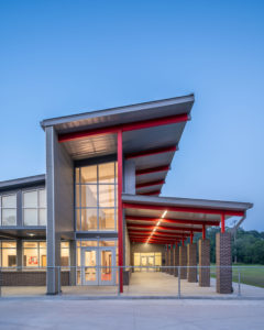 A modern school building entrance at dusk, featuring a distinctive architectural design with a long, slanted metal roof, red structural beams, large windows, and an illuminated covered walkway with brick columns.