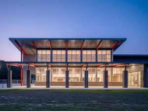 A modern building facade at dusk, featuring a distinctive, large, overhanging metal roof supported by red and gray columns. The two-story structure has large windows on both levels and is brightly lit from within.