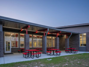 A modern school building patio at dusk, featuring a covered outdoor eating area with red picnic tables. The space has a brick and gray facade, large windows, and an overhanging roof supported by red and dark gray columns.