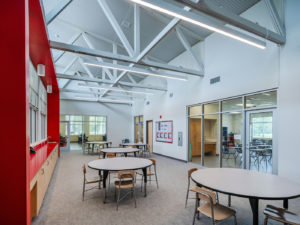 A wide-angle view of a modern school multi-purpose room with a high, exposed truss ceiling and large windows. The space features several round tables and chairs, a bold red accent wall with a built-in counter and sink, and a view into an adjacent classroom.