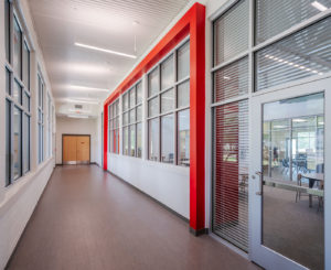 A modern school hallway with polished floors, white walls, and a distinctive red architectural frame surrounding a long row of large windows. The windows provide a view into a classroom, and a set of double doors is visible at the end of the hall.
