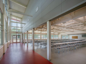 A spacious school cafeteria with numerous long tables and benches arranged in rows. The room features high ceilings with exposed wooden trusses, large windows for natural light, and a contrasting red and gray floor.