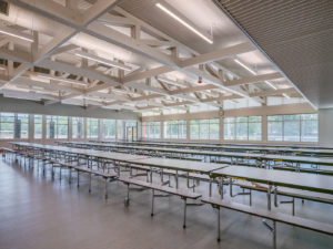 A spacious school cafeteria with numerous long tables and benches arranged in rows. The room features high ceilings with exposed wooden trusses, large windows for natural light, and a contrasting red and gray floor.