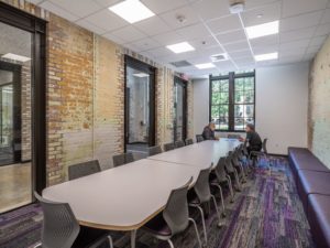 A modern conference room features a long table with numerous chairs, exposed brick walls, and large windows, with two people seated at the far end of the table.