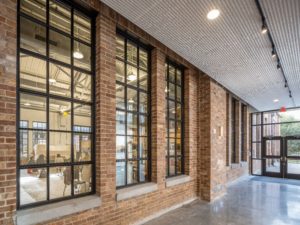 A wide hallway with exposed brick walls, a polished concrete floor, and a row of large, multi-paned windows looking into a bright, open studio or classroom space.