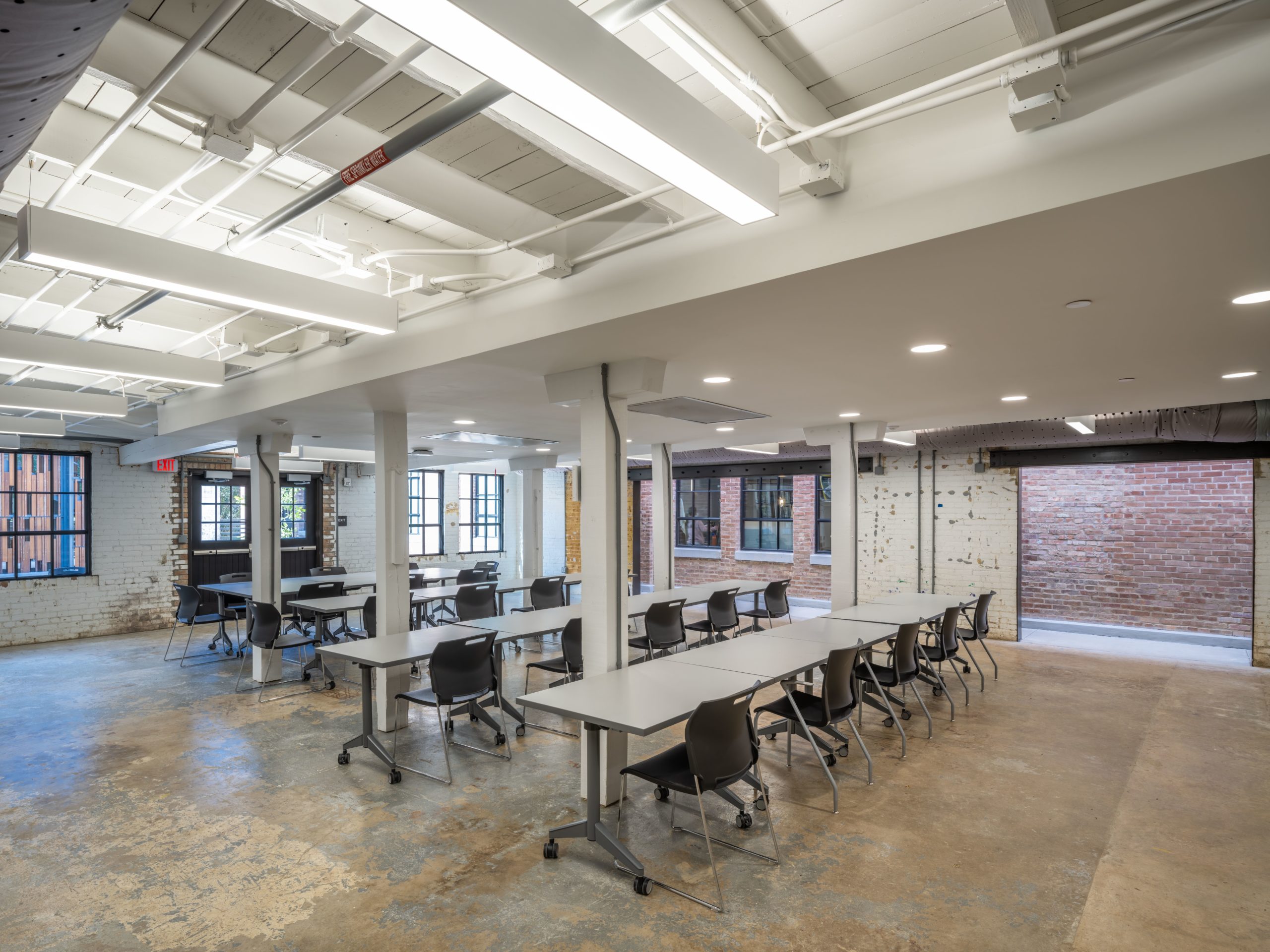 A wide shot of a modern, industrial-style classroom or workshop space with multiple long tables and chairs arranged in rows. The room features polished concrete floors, exposed ceiling pipes, large windows, and brick walls.
