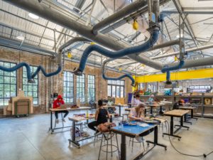 A busy workshop or makerspace with a high ceiling, exposed brick walls, and large windows. Multiple people are seated at workbenches and tables, engaged in various projects under a system of industrial ventilation pipes.