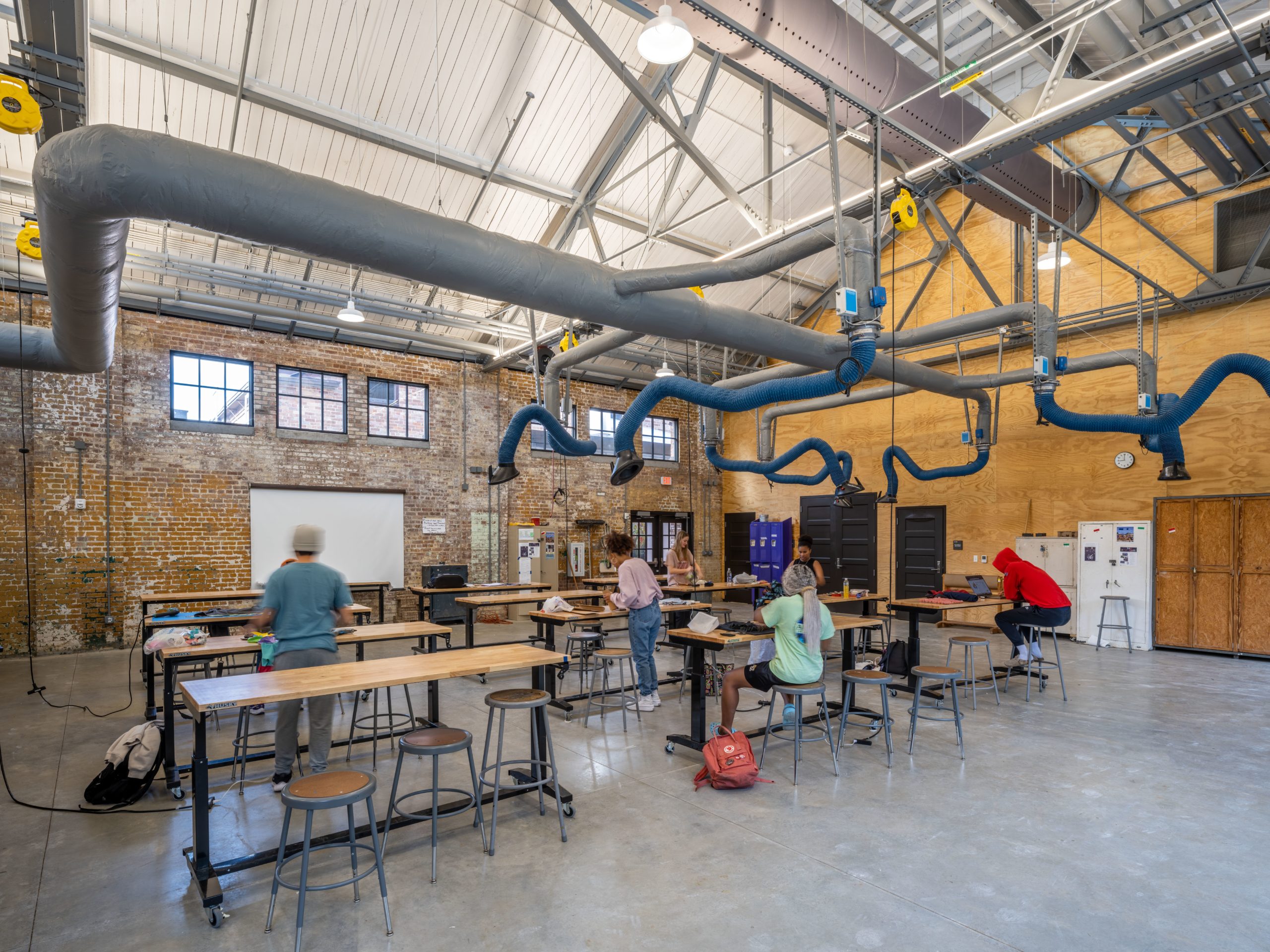 A bright and open workshop or makerspace with a high ceiling, exposed brick walls, and visible ductwork. Several people are working at long tables, engaged in creative projects, with a whiteboard in the background.