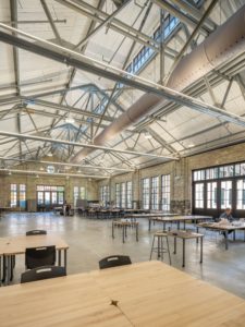A spacious, modern classroom or studio with a high, industrial-style ceiling, exposed metal trusses, and concrete floors. The room is filled with multiple worktables, chairs, and large windows, with a person seated at a desk in the background.